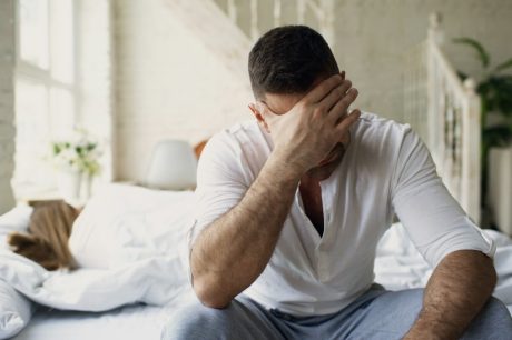 Upset young man sitting in bed have problems with sex while his girlfriend sleep in bedroom in the morning